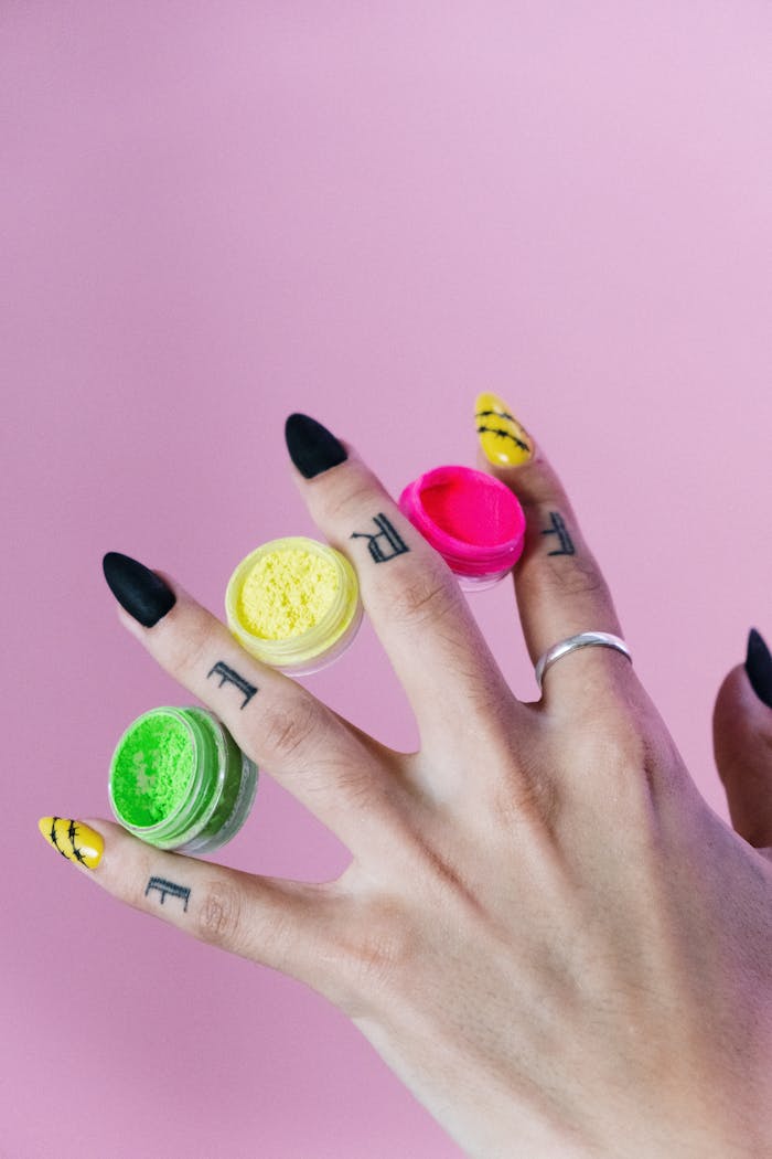 Close-up of a tattooed hand with colorful nail polish jars on fingers against a pink background.